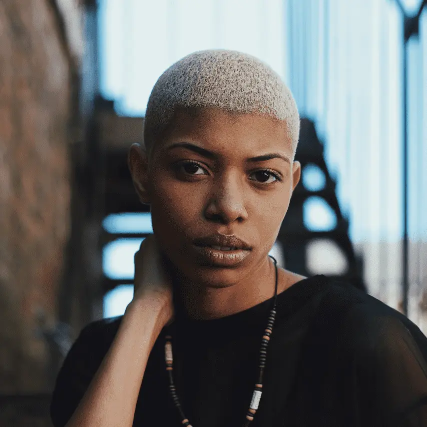 A close-up portrait of a person with short, buzzed blonde hair and a thoughtful expression. They are wearing a black top and a beaded necklace, posing with one hand resting against their neck. The background shows a soft-focus industrial setting with stairs.