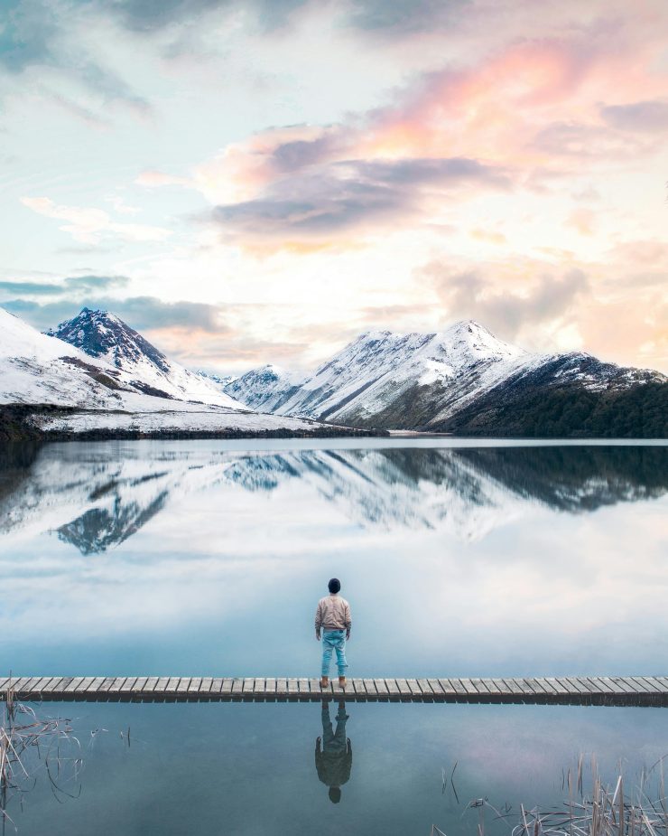A person standing on a wooden dock overlooking a calm lake surrounded by snow-capped mountains under a pastel sky.