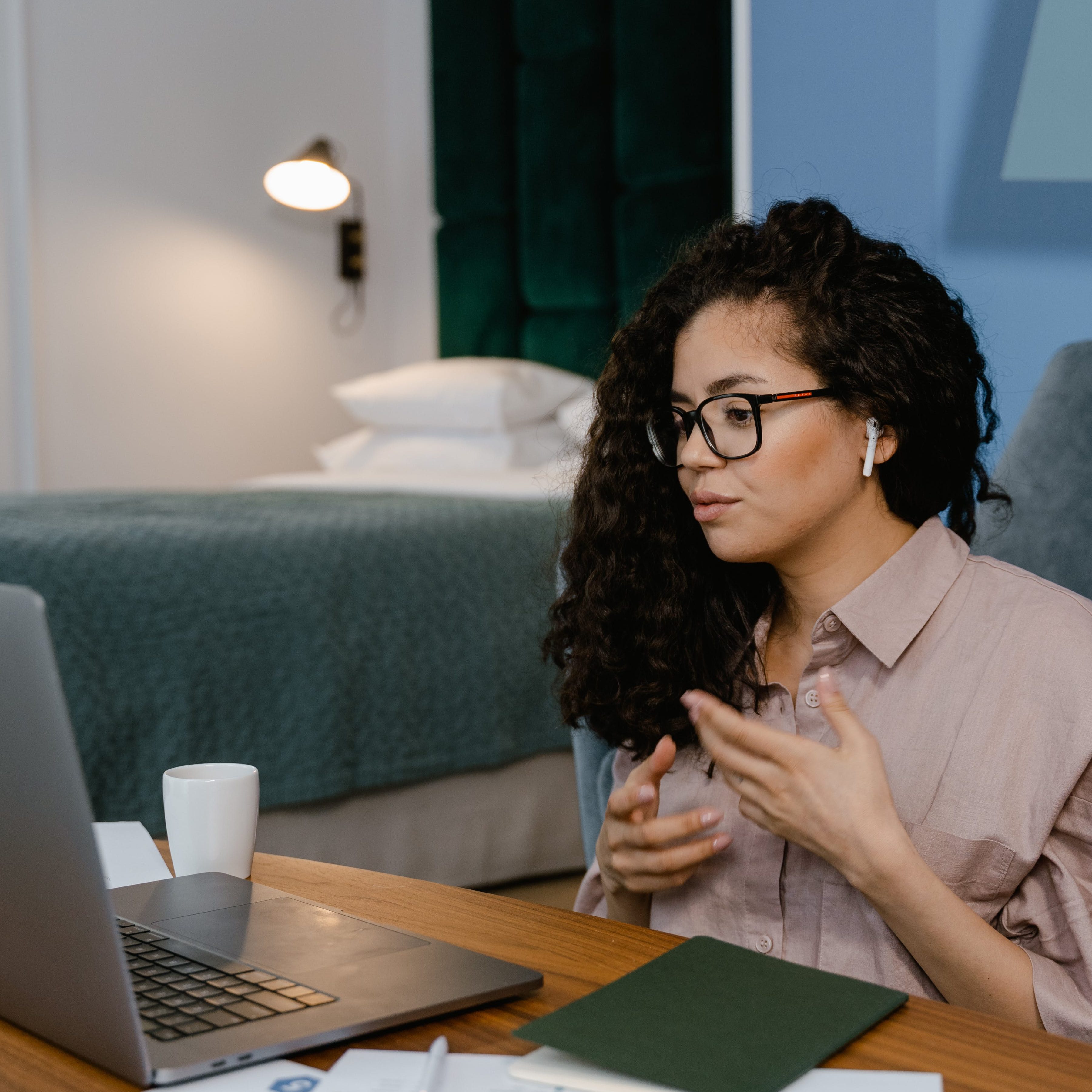 A woman with long, curly dark hair and glasses sitting at a wooden desk, gesturing with her hands while looking at a laptop. She is wearing a light pink button-down shirt and wireless earbuds. In the background, a neatly made bed and a green headboard are visible in a brightly lit room.